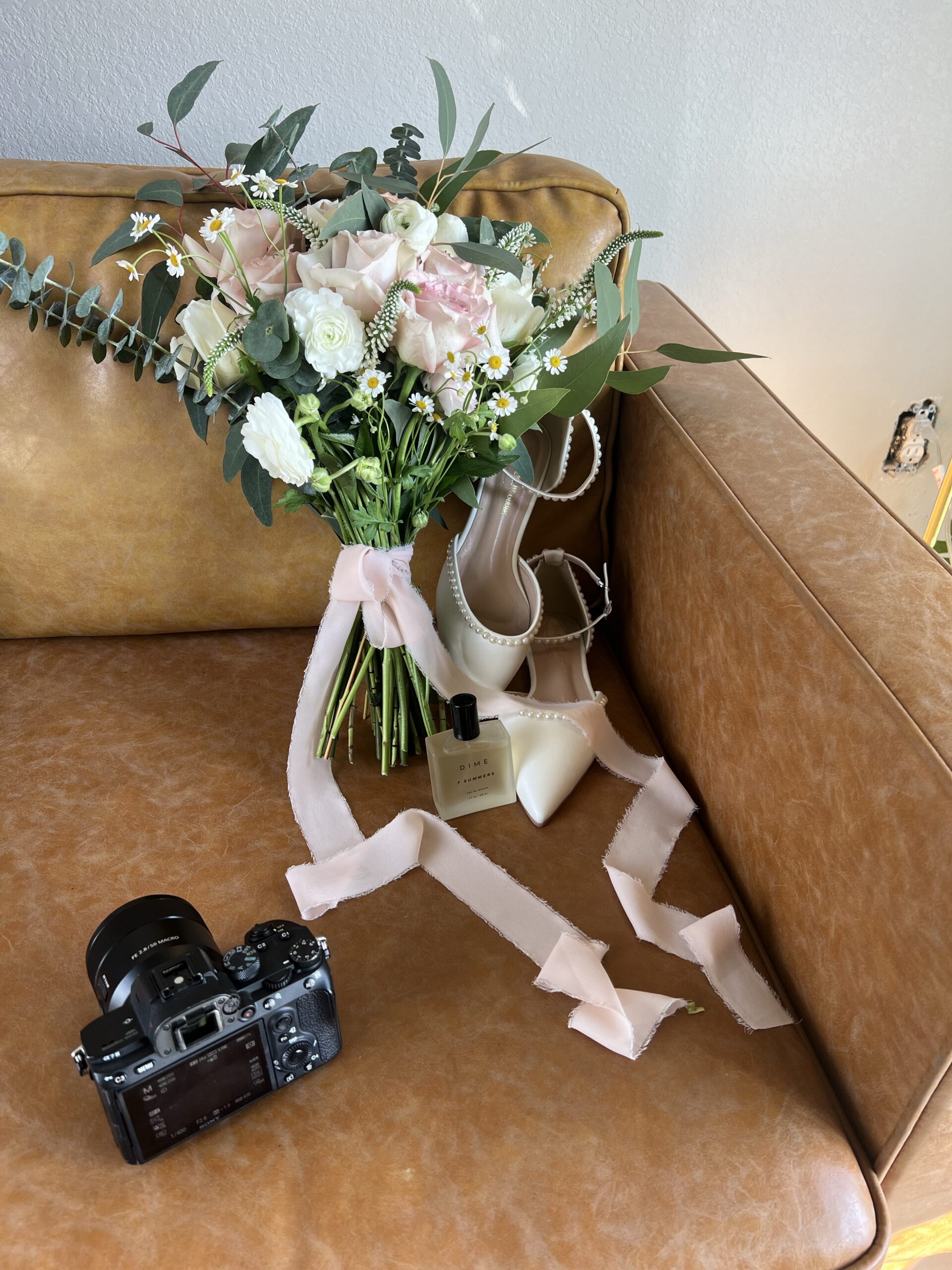 A wedding bouquet with pink ribbon sitting next to a camera on a leather brown couch