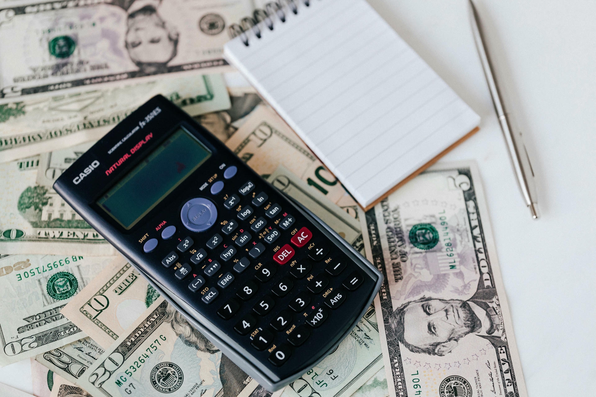 Calculator resting on scattered dollar bills next to a notepad and pen, wedding budget planning concept
