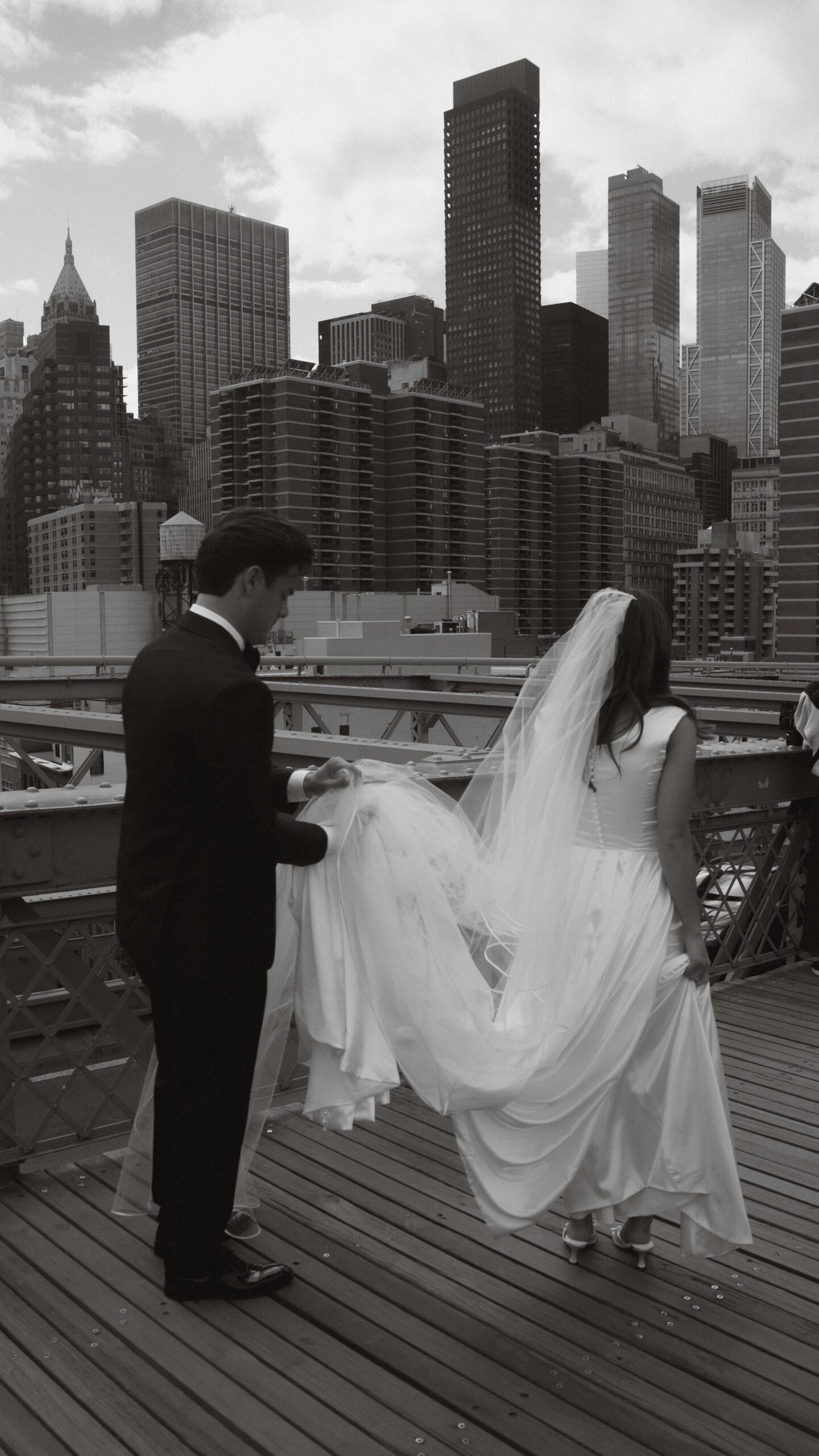 Groom holding the bride's flowing veil on a wooden bridge, black and white wedding portrait.