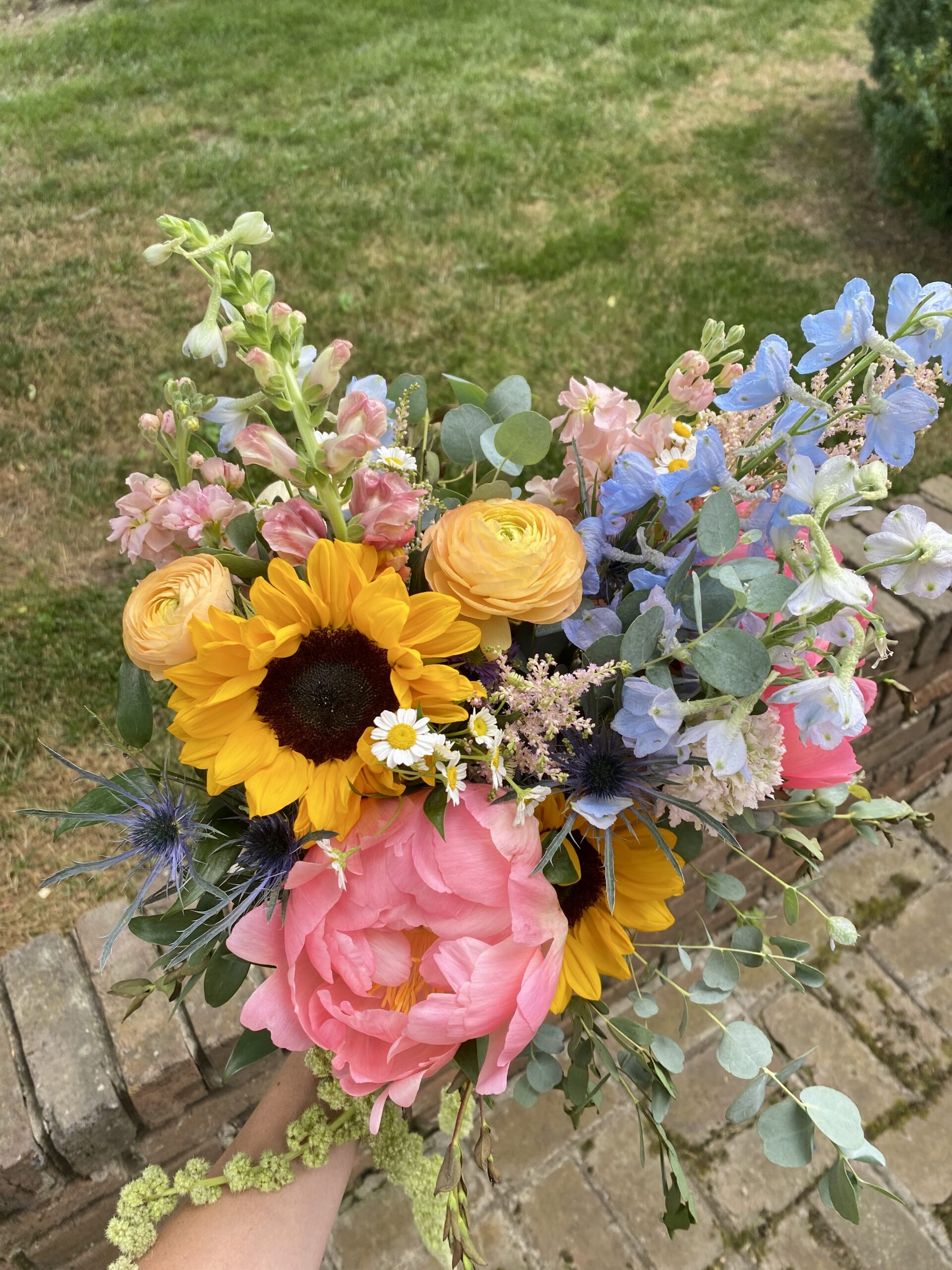 A hand holds a bright wedding bouquet with sunflowers, pink peonies, yellow ranunculus, blue delphinium, and eucalyptus greenery, against a grassy backyard and brick patio.